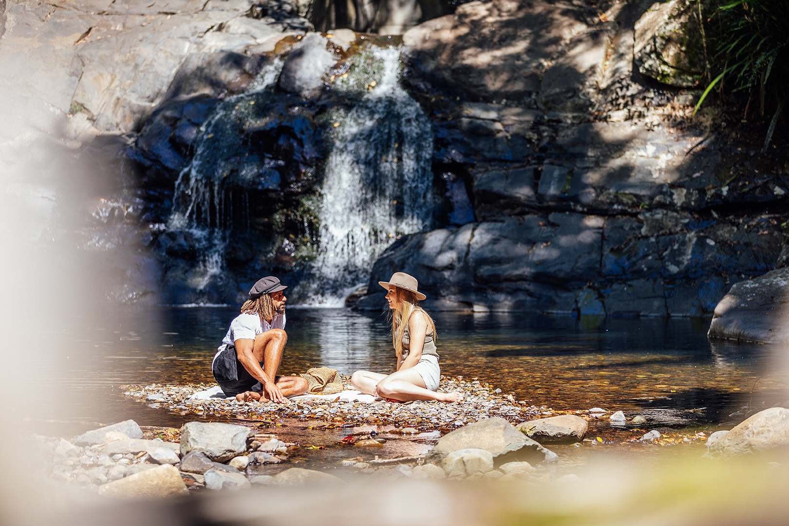 A young couple sitting on the rocks along the shores of the rock pools at Cougal Cascades. A waterfall is in the background.