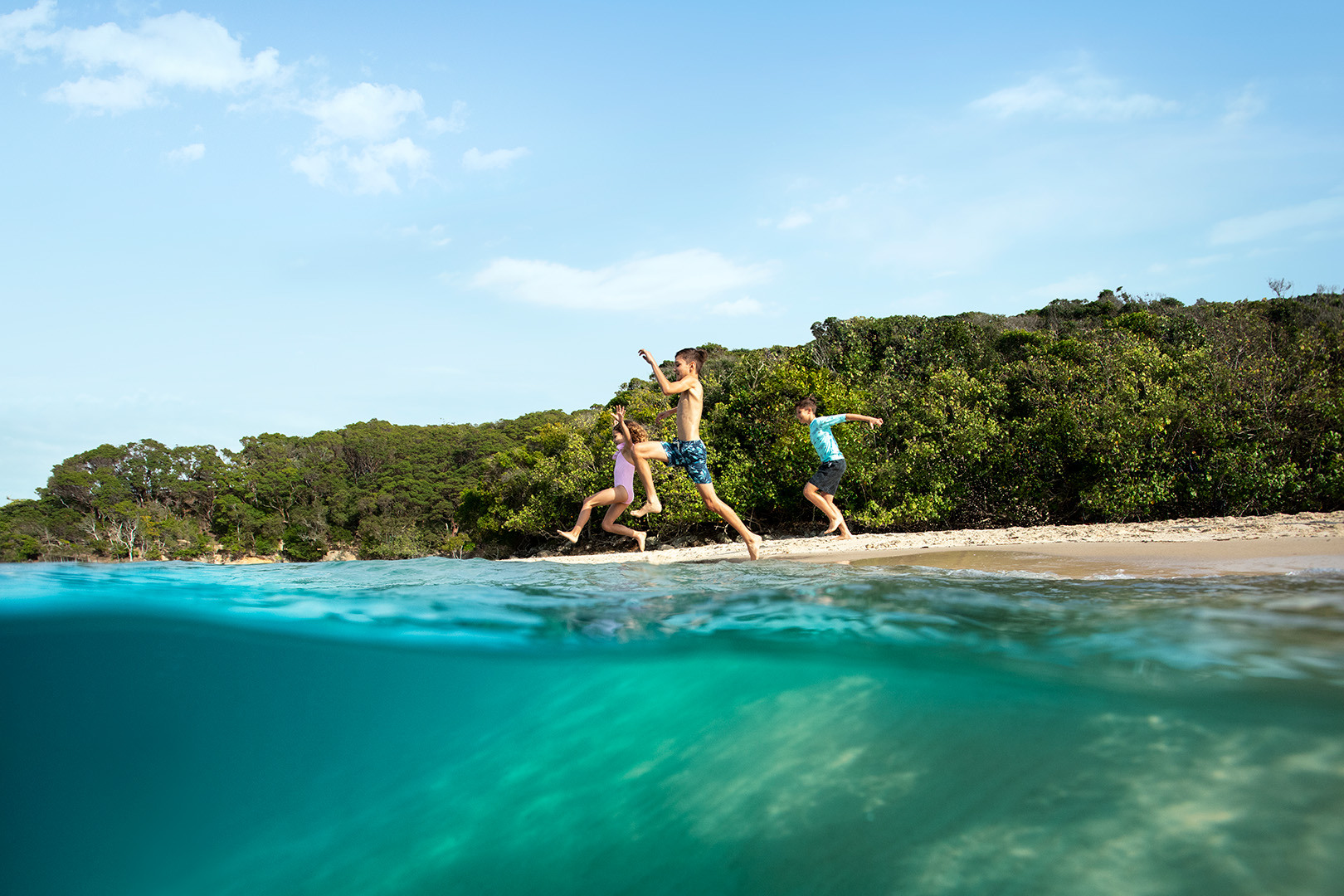 Three kids jumping from the shore into Tallebudgera Creek on a sunny day.