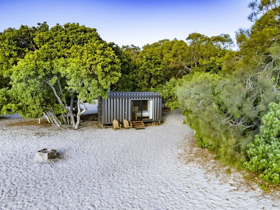 A small, modern cabin made from a shipping container sits on a sandy beach surrounded by lush green trees, featuring a wooden deck with two wicker chairs and a table, a nearby stone fire pit, and a clear blue sky overhead.